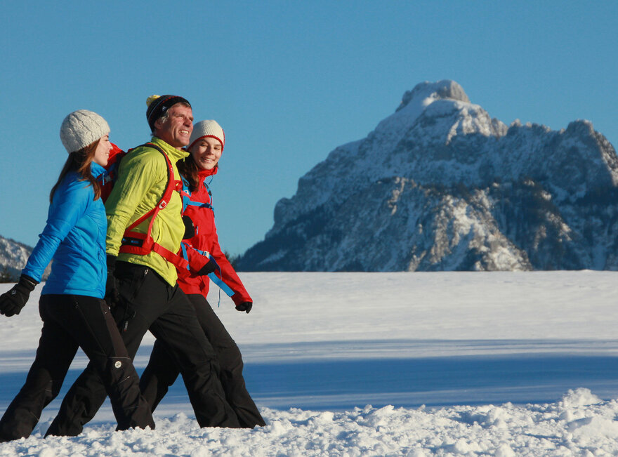 Das Bild zeigt zwei Frauen und einen Mann. Sie tragen Sport-Winterkleidung und laufen nebeneinander auf einem verschneiten Weg. Alle lächeln. Im Hintergrund sieht man die schneebedeckte Umgebung und die verschneiten Berge. Der Himmel ist blau. Die Sonne scheint.  