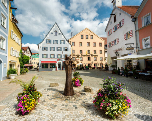 Das Bild zeigt eine gemütliche mittelalterliche Stadt mit farbenfrohen Gebäuden, Kopfsteinpflasterstraßen und Blumenkübeln. Die Stadt ist lebhaft mit Menschen befüllt und es gibt mehrere Sonnenschirme und Tische für eine gemütliche Einkehr.