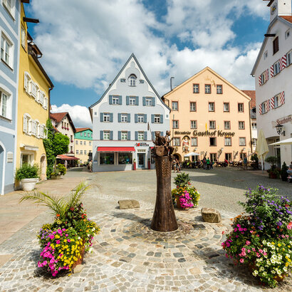 Das Bild zeigt eine gemütliche mittelalterliche Stadt mit farbenfrohen Gebäuden, Kopfsteinpflasterstraßen und Blumenkübeln. Die Stadt ist lebhaft mit Menschen befüllt und es gibt mehrere Sonnenschirme und Tische für eine gemütliche Einkehr.