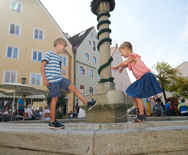 Das Bild zeigt zwei Kinder, die in einer Springbrunnenanlage spielen. Die Kinder stehen am Rand der Springbrunnenanlage, und eines der Kinder spitzt mit dem Wasser.