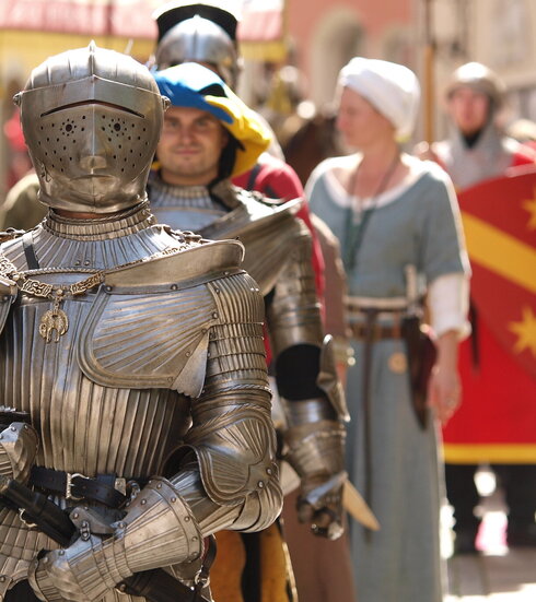 Das Bild erfasst eine lebhafte Szene eines mittelalterlich-thematisierten Festes. Das Hauptaugenmerk liegt auf einem Mann in einer vollständigen Rüstung, inklusive Helm und Schwert. Er steht in der Mitte des Bildes und zieht die Aufmerksamkeit des Betrachters auf sich. Zu seiner Linken befindet sich eine Frau in mittelalterlichem Gewand und hält einen Schild. Zu seiner Rechten steht ein weiterer Mann in Rüstung, der ein Banner hält. Im Hintergrund sind andere Personen in mittelalterlichem Gewand zu sehen, die zur festlichen Stimmung beitragen. Die Gebäude im Hintergrund sowie die Kostüme zeigen die mittelalterlichen Häuser der Altstadt von Füssen. Die gesamte Szene ist ein lebhafter Ausdruck mittelalterlicher Kultur und Geschichte.