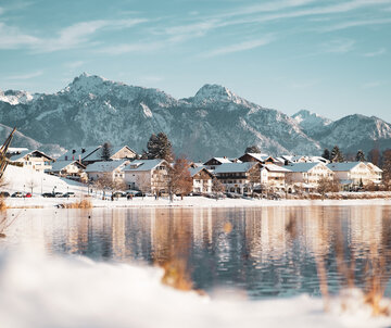 Das Bild zeigt eine verschneite Winterszenerie. Schneebedeckte Häuser stehen am Ufer eines Sees. Im Hintergrund erheben sich die ebenfalls schneebedeckten Berge. Der Himmel ist blau. Die Sonne scheint. Die Wasseroberfläche des Sees ist ruhig.  