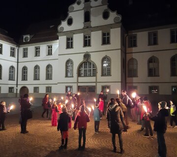 Das Bild zeigt eine große Gruppe von Erwachsenen und Kindern, die sich im Innenhof des Hohen Schloss in Füssen versammelt haben und Fackeln halten, die brennen. Die Menge wird durch das Leuchten der Fackeln erhellt, was eine warme und einladende Atmosphäre schafft.