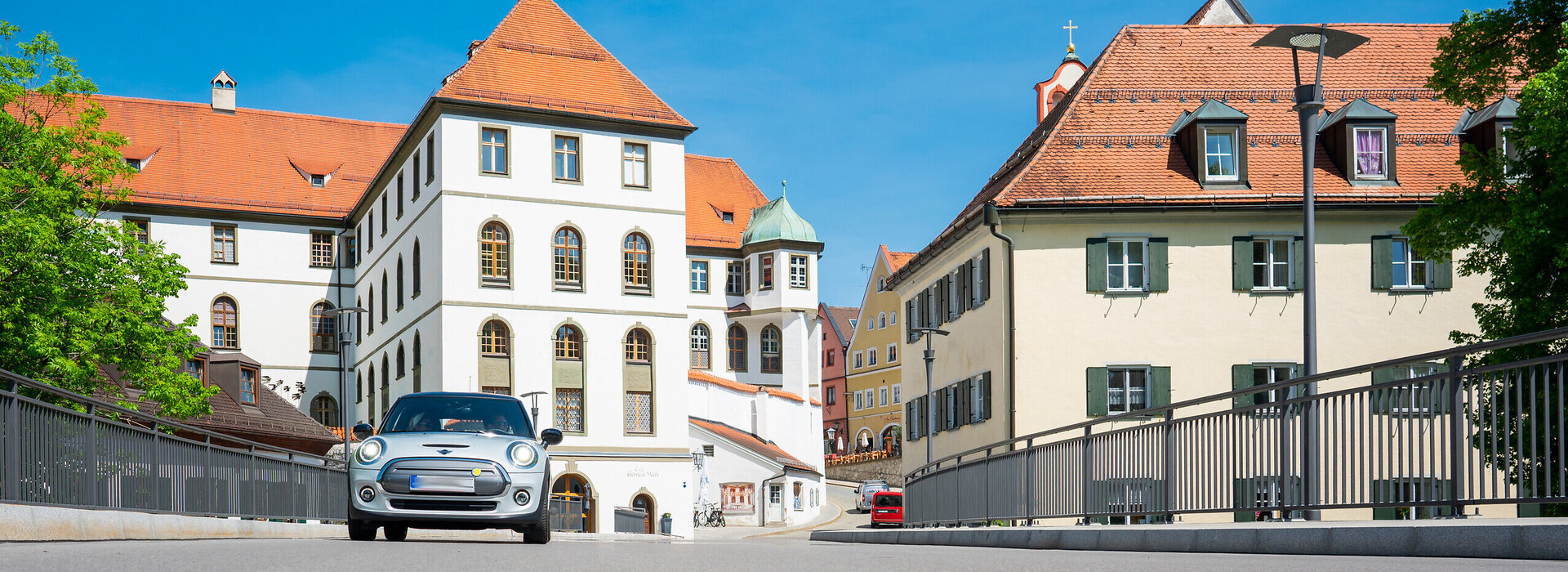 Ein silberner Mini fährt auf den Betrachter zu. Er fährt über eine Brücke. Im Hintergrund erhebt sich neben den bunten Altstadthäusern, der Gebäudekomplex des ehemaligen Benediktinerklosters mit seinen roten Dächern und unzähligen Fenstern. Der Himmel ist blau. Die Sonne scheint. 