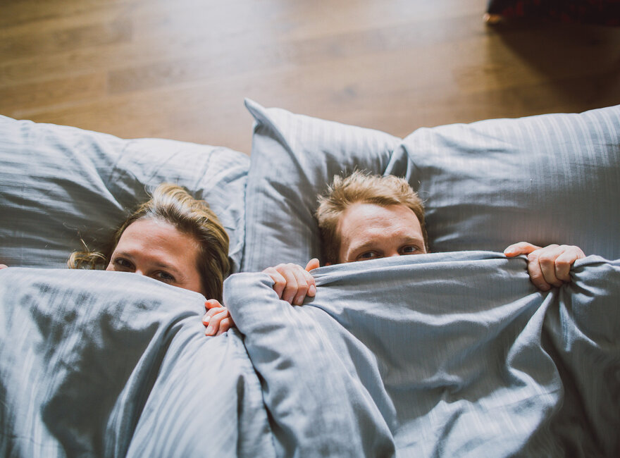 Das Bild zeigt eine Frau und einen Mann von oben, die sich eine blaugraue Bettdecke über die Nasenspitze ziehen und in die Kamera blicken. Ihre Köpfe sind auf ebensoblaugrauen Kissen gebettet.