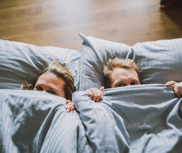 Das Bild zeigt eine Frau und einen Mann von oben, die sich eine blaugraue Bettdecke über die Nasenspitze ziehen und in die Kamera blicken. Ihre Köpfe sind auf ebensoblaugrauen Kissen gebettet.