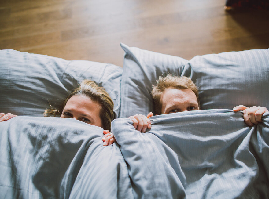 Das Bild zeigt eine Frau und einen Mann von oben, die sich eine blaugraue Bettdecke über die Nasenspitze ziehen und in die Kamera blicken. Ihre Köpfe sind auf ebensoblaugrauen Kissen gebettet.