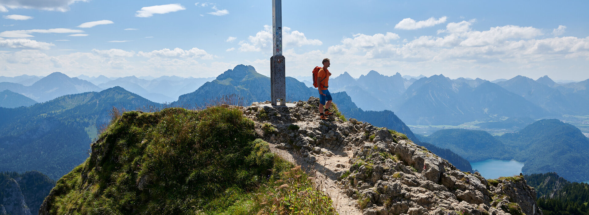 Das Bild zeigt einen Mann, der auf einem Berggipfel steht, wo ein Kreuz darauf ist. lt. Der Mann trägt einen Rucksack und blickt über eine gebirgige Landschaft. Im Hintergrund kann man einen See und Berge erkennen.