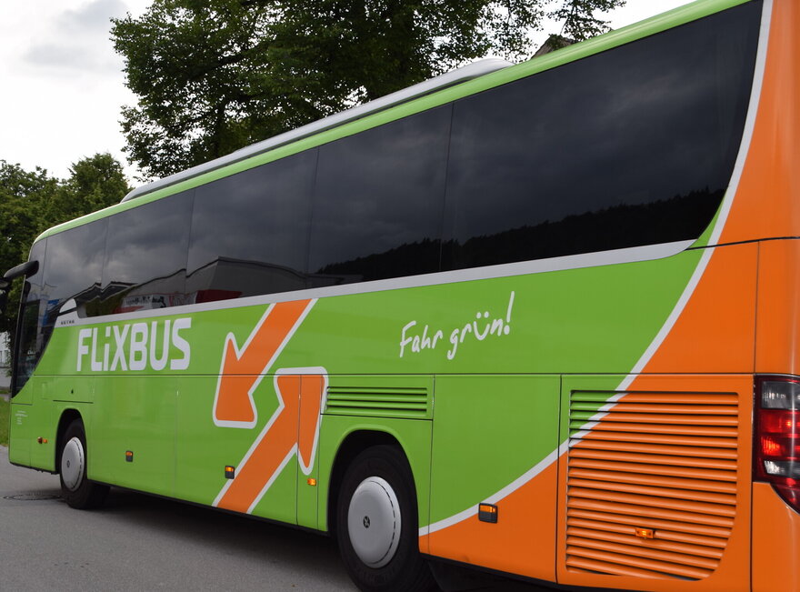 Im Vordergrund ist ein grüner, orangener Flixbus zu sehen. Der am Straßenrand steht. Der Himmel ist wolkenverhangen.  