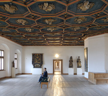 Man blickt in einen Saal mit weißen Wänden, einem historischen Holzboden und einer reich verzierten, dunkelblauen Holzdecke. Das Muster der Decke ist wabenförmig. Die Kanten der Waben sind erhaben und mit roten Umrandungen versehen. Innerhalb jeder Wabe ist eine, in Gold gearbeitete, kirchliche Person zu sehen. Man sieht Heiligenfiguren, aber auch Bischöfe und Päpste. An den Wänden hängen historische Gemälde mit unterschiedlichen Themen. Der Raum ist, bis auf eine moderne mit schwarzem Leder bezogene Bank in der Mitte, nicht möbliert. Auf der Bank sitzt eine Frau und blickt auf die Gemälde.