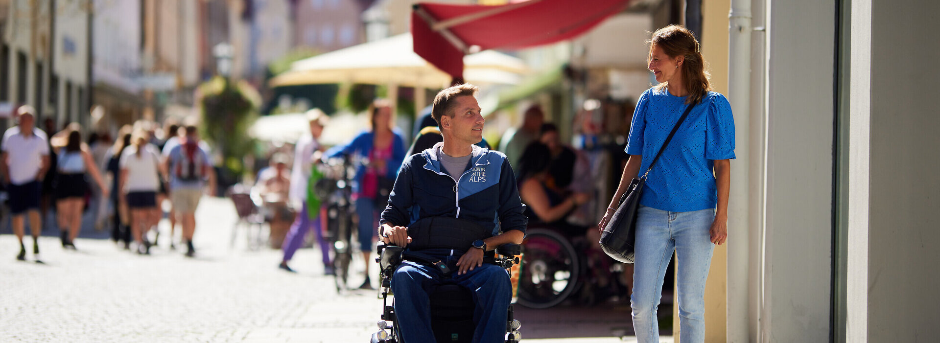  Das Bild zeigt eine lebhafte Straßenszene in einer europäischen Stadt. Der Blickfang des Bildes ist ein Mann im Rollstuhl, der sich in einem Gespräch mit einer Frau befindet, die neben ihm geht. Der Mann ist mit einem blauen Jacket und einem schwarzen Hut gekleidet, während die Frau ein blaues Hemd und blaue Jeans trägt. Sie gehen auf einem Gehweg, der mit unterschiedlichen Geschäften und Restaurants gesäumt ist. Die Straße ist belebt mit Menschen, von denen einige zu Fuß gehen, während andere Fahrräder fahren. Die Gebäude im Hintergrund sind hoch und haben einen europäischen architektonischen Stil. Der Himmel über ihnen ist klar und erlaubt den Schluss auf einen angenehmen Tag. Die Gesamtatmosphäre des Bildes ist lebhaft und voller Leben.