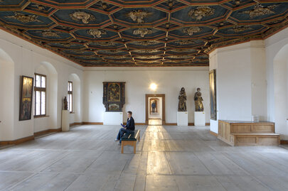 Man blickt in einen Saal mit weißen Wänden, einem historischen Holzboden und einer reich verzierten, dunkelblauen Holzdecke. Das Muster der Decke ist wabenförmig. Die Kanten der Waben sind erhaben und mit roten Umrandungen versehen. Innerhalb jeder Wabe ist eine, in Gold gearbeitete, kirchliche Person zu sehen. Man sieht Heiligenfiguren, aber auch Bischöfe und Päpste. An den Wänden hängen historische Gemälde mit unterschiedlichen Themen. Der Raum ist, bis auf eine moderne mit schwarzem Leder bezogene Bank in der Mitte, nicht möbliert. Auf der Bank sitzt eine Frau und blickt auf die Gemälde. 