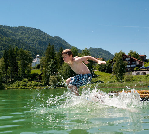Das Bild zeigt einen Jungen, der in den Weißensee springt. Er hat die Wasseroberfläche bereits durchbrochen und das Wasser spritzt in alle Richtungen umher. Der Junge trägt blaue Badehosen. Der Weißensee ist von grünen Bäumen und Hügeln umgeben. Der Himmel ist blau. Die Sonne scheint.  
