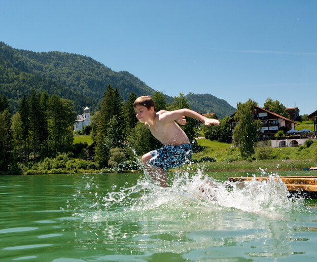 Das Bild zeigt einen Jungen, der in den Weißensee springt. Er hat die Wasseroberfläche bereits durchbrochen und das Wasser spritzt in alle Richtungen umher. Der Junge trägt blaue Badehosen. Der Weißensee ist von grünen Bäumen und Hügeln umgeben. Der Himmel ist blau. Die Sonne scheint.  