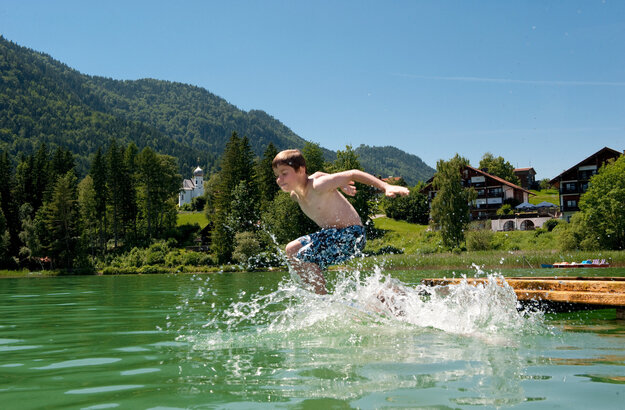 Das Bild zeigt einen Jungen, der in den Weißensee springt. Er hat die Wasseroberfläche bereits durchbrochen und das Wasser spritzt in alle Richtungen umher. Der Junge trägt blaue Badehosen. Der Weißensee ist von grünen Bäumen und Hügeln umgeben. Der Himmel ist blau. Die Sonne scheint.  