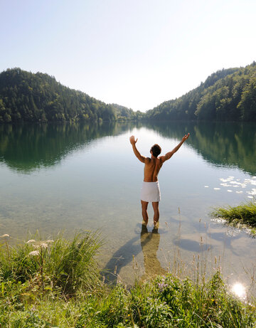Das Bild zeigt einen Mann, der sich nur ein weißen Handtuch um die Hüften gebunden hat. Er hat dem Betrachter den Rücken zugedreht, steht im knöcheltiefen Wasser eines Sees und streckt die Arme nach oben. Der See ist umringt von grünen Wäldern. Der Himmel ist blau. Die Sonne scheint.        