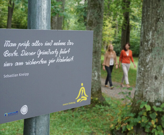 Das Bild zeigt zwei Frauen, die auf einem Waldweg laufen. Ringsherum wachsen hohe Bäume. Auf dem Schild im Vordergrund steht „Man prüfe alles und nehme das Beste. Dieser Grundsatz führt uns am sichersten zur Wahrheit. Sebastian Kneipp“