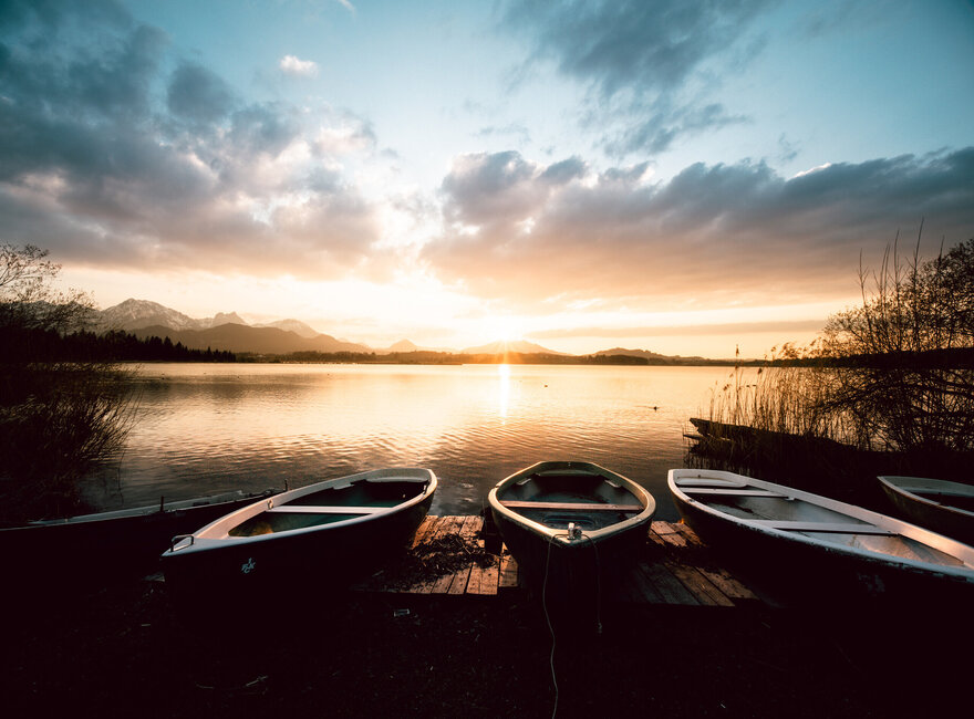 Das Bild zeigt einen Sonnenuntergang am Ufer des Hopfensees. Der Himmel ist in orange und blau gefärbt und dient als beeindruckende Kulisse für den ruhigen See. Am Ufer des Sees liegen, im Zentrum des Bildes drei Ruderboote an Land. Die Ruderboote, die in Größe und Verarbeitung unterschiedlich sind, sind leer und warten still auf ihr nächstes Abenteuer. 