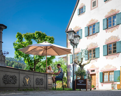 Das Foto zeigt einen Mann und eine Frau, die an einem Tisch unter einem Sonnenschirm sitzen und eine Speisekarte lesen. Im Hintergrund erhebt sich eine dreistöckiges Haus mit einem Spitzen Giebel. Seine Fenster sind von grünen Fensterläden umrahmt. Im Vordergrund ist ein dunkelgrauer, wohl gusseiserner Brunnen zu sehen. Der Himmel ist blau. Die Sonne scheint.