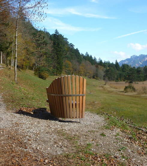 Man blickt über eine hölzerne Bank in ein Tal. Auf einer Anhöhe verläuft ein nicht befestigter Waldweg, der von Bäumen gesäumt ist. Unterhalb des Wegs liegt eine Wiese, an die ein Schilfgürtel angrenzt. Im Hintergrund erheben sich die Berge. Davor ist ein Wald aus Nadelbäumen zu sehen. 