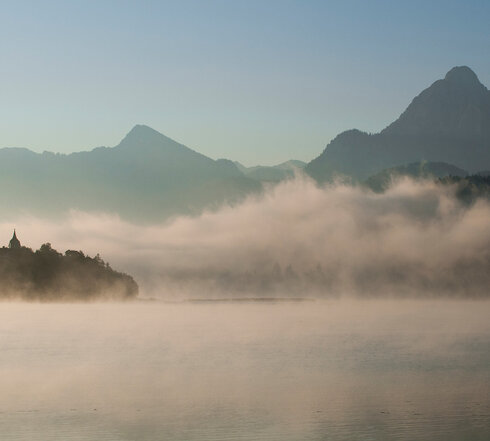 Das Bild zeigt in Nebel gehüllte Berge, die sich majestätisch im Hintergrund erheben.  Der zartweiße Nebel verleiht der Szene einen Hauch von mystischer Stimmung. Im Vordergrund spiegelt der ruhige Weißensee die majestätischen Berge. Über den tiefblauen See ziehen die feinen Nebelschwaden hinweg. Der Himmel darüber ist hell und blau. 