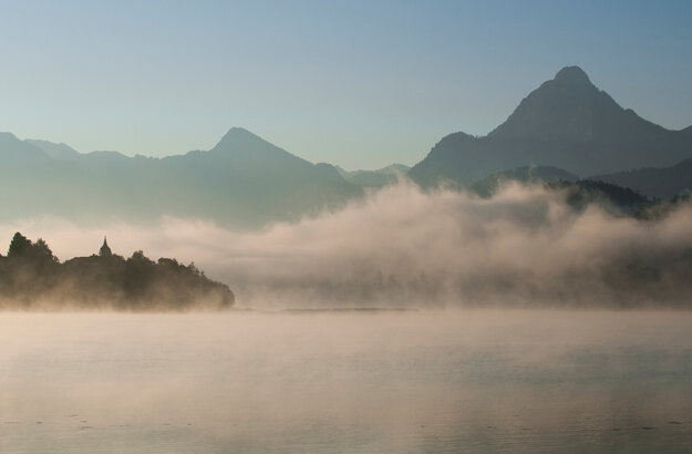 Das Bild zeigt in Nebel gehüllte Berge, die sich majestätisch im Hintergrund erheben.  Der zartweiße Nebel verleiht der Szene einen Hauch von mystischer Stimmung. Im Vordergrund spiegelt der ruhige Weißensee die majestätischen Berge. Über den tiefblauen See ziehen die feinen Nebelschwaden hinweg. Der Himmel darüber ist hell und blau. 
