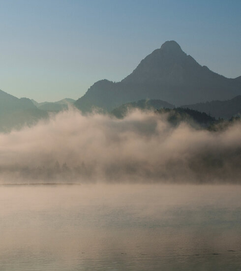 Das Bild zeigt in Nebel gehüllte Berge, die sich majestätisch im Hintergrund erheben.  Der zartweiße Nebel verleiht der Szene einen Hauch von mystischer Stimmung. Im Vordergrund spiegelt der ruhige Weißensee die majestätischen Berge. Über den tiefblauen See ziehen die feinen Nebelschwaden hinweg. Der Himmel darüber ist hell und blau. 