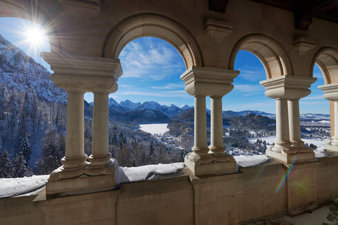 Das Bild fängt einen atemberaubenden Blick auf eine schneebedeckte Bergkette ein, die vom hohen Aussichtspunkt aus betrachtet wird. Die Perspektive ist von einem Balkon aus, der mit einer Reihe von Bögen geschmückt ist. Diese Bögen, aus Stein gefertigt, sind gleichmäßig gesetzt und verleihen der Szene ein Gefühl von Symmetrie und Gleichgewicht. Die Sonne scheint hell, wirft ein warmes Glühen auf die schneebedeckten Berge und beleuchtet die Bögen so, dass ihre architektonische Schönheit betont wird.