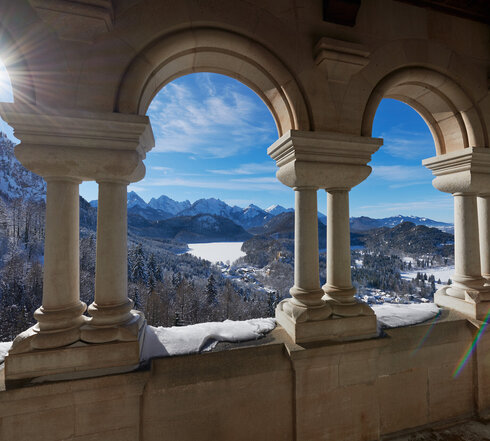 Das Bild fängt einen atemberaubenden Blick auf eine schneebedeckte Bergkette ein, die vom hohen Aussichtspunkt aus betrachtet wird. Die Perspektive ist von einem Balkon aus, der mit einer Reihe von Bögen geschmückt ist. Diese Bögen, aus Stein gefertigt, sind gleichmäßig gesetzt und verleihen der Szene ein Gefühl von Symmetrie und Gleichgewicht. Die Sonne scheint hell, wirft ein warmes Glühen auf die schneebedeckten Berge und beleuchtet die Bögen so, dass ihre architektonische Schönheit betont wird.