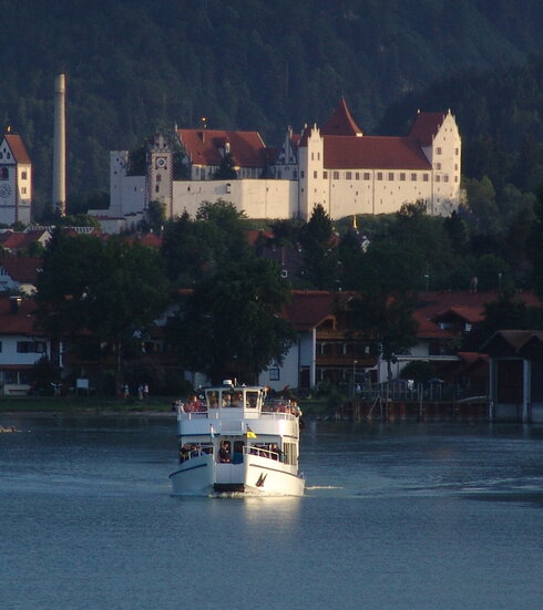 Das Bild zeigt ein zweistöckiges Schiff, das auf einem See auf den Betrachter zufährt. Im Hintergrund thront auf eine Anhöhe das Hohe Schloss von Füssen mit seinen Türmen, Mauern und Fenstern und unterschiedlichen Gebäudeteilen. Dahinter erhebt sich ein bewaldeter Berg. Die Sonne scheint. 