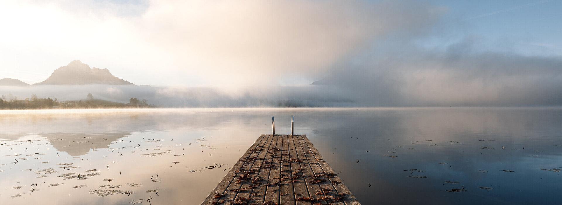 Das Bild zeigt einen Holzsteg, der auf einen See führt. Auf dem Steg liegt buntes Laub. Am Ende des Stegs führt eine Leiter ins Wasser. Die Wasseroberfläche des Sees ist ruhig und fast wie ein Spiegel. Im Hintergrund erheben sich Berge aus dem Nebel. Über dem Nebel ist der blaue Himmel zu sehen. Die Sonne bahnt sich scheinbar ihren Weg.  