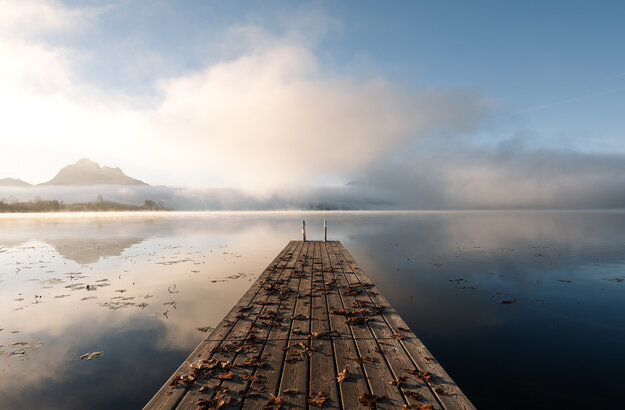 Das Bild zeigt einen Holzsteg, der auf einen See führt. Auf dem Steg liegt buntes Laub. Am Ende des Stegs führt eine Leiter ins Wasser. Die Wasseroberfläche des Sees ist ruhig und fast wie ein Spiegel. Im Hintergrund erheben sich Berge aus dem Nebel. Über dem Nebel ist der blaue Himmel zu sehen. Die Sonne bahnt sich scheinbar ihren Weg.  