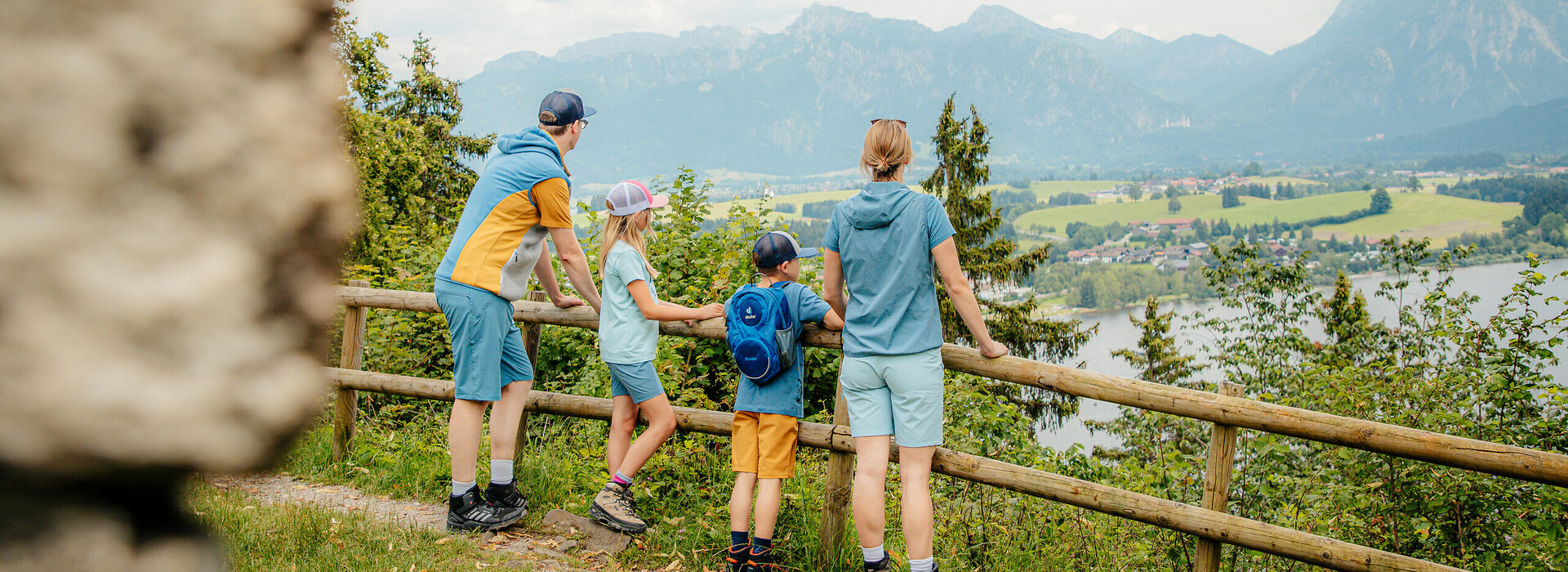 Das Bild zeigt einen idyllischen Moment in einer gebirgigen Landschaft. Eine Familie von vier Personen steht an einem hölzernen Zaun. Die Eltern sind im Bild in blauen Hemden und kurzen Hosen gekleidet, die Kinder in orangen Hemden und kurzen Hosen. Sie alle blicken in die gleiche Richtung, ihre Blicke sind auf den atemberaubenden Anblick der Berge in der Ferne gerichtet. Der Himmel über ihnen ist ein strahlendes Blau, durchzogen von flauschigen weißen Wolken. Die Familie scheint die Zeit miteinander zu genießen und die Schönheit der Natur zu schätzen. Der hölzerne Zaun, an dem sie stehen, verleiht der Szene einen rustikalen Charme. Die allgemeine Atmosphäre des Bildes ist eine von Ruhe und familiärer Verbundenheit.