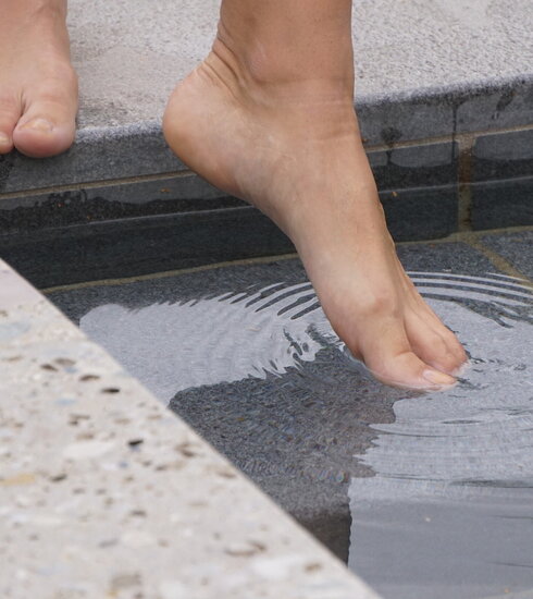 Das Bild eine Person, die vor einem Tretbecken steht und den linken Fuß ins Wasser streckt. Allerdings sind nur die nackten Füße der Person zu sehen. 