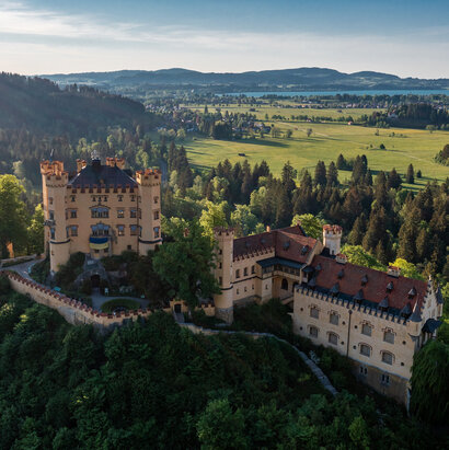 Das Bild zeigt das Schloss Hohenschwangau, das auf einem Hügel thront und von üppigem grünen Wald umgeben ist. Die Burg, aus Stein erbaut, verfügt über mehrere Türme und Erker, welche zu ihrer Größe beitragen. Die Architektur der Burg erinnert an eine mittelalterliche Festung, ausgestattet mit Wehrgängen und einem Graben. Die umliegende Landschaft ist gesprenkelt mit grünen Bäumen und sanften Hügeln, was eine malerische Kulisse schafft.
