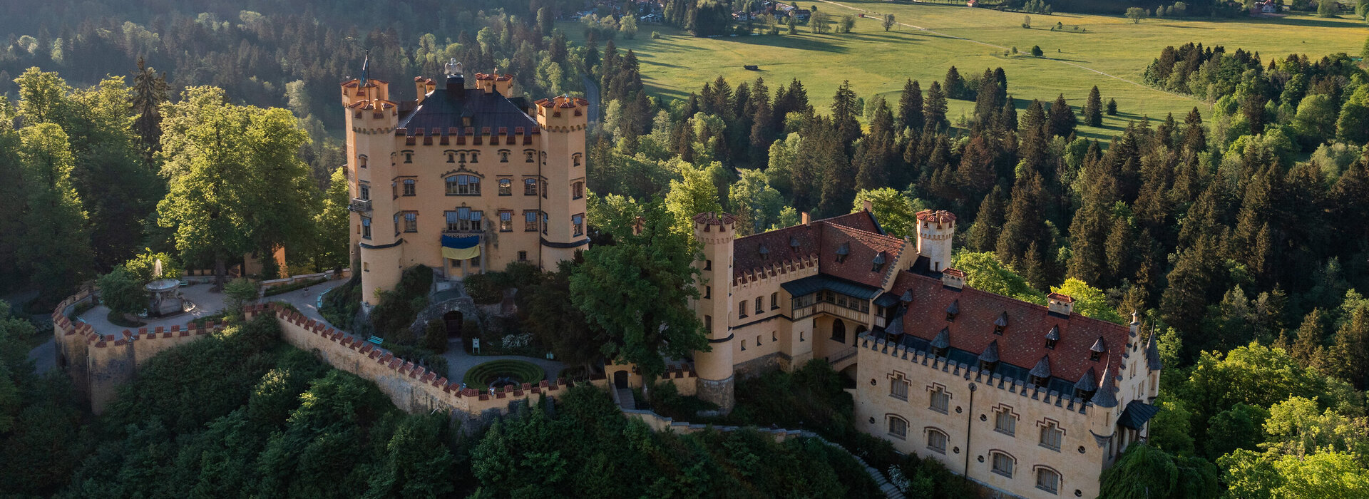 Das Bild zeigt das Schloss Hohenschwangau, das auf einem Hügel thront und von üppigem grünen Wald umgeben ist. Die Burg, aus Stein erbaut, verfügt über mehrere Türme und Erker, welche zu ihrer Größe beitragen. Die Architektur der Burg erinnert an eine mittelalterliche Festung, ausgestattet mit Wehrgängen und einem Graben. Die umliegende Landschaft ist gesprenkelt mit grünen Bäumen und sanften Hügeln, was eine malerische Kulisse schafft.