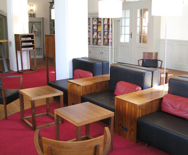 Man blickt in einen hohen Raum mit Bogenfenster. Im Vordergrund stehen schwarze Ledersessel mit Holzlehnen und niedrigen Holztischen. Auf den Ledersesseln liegen rote Kissen. Auf dem Holzdielenboden liegen rote, runde Teppiche. Im Hintergrund sind Vitrinen mit Zeitschriften zu sehen. Der Raum ist hell und einladend.