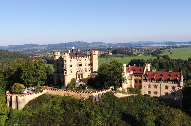 Das Bild zeigt das gelbe Schloss Hohenschwangau, dass auf einer Anhöhe thront und von Bäumen umgeben ist. Das Schloss besteht aus zwei Gebäudeteilen. Der quadratische, vierstöckige Hauptbau ist an jeder Ecke mit einem Turm versehen, der bis zum Dach reicht. Das längliche Nebengebäude steht mit ein bisschen Abstand zum Hauptgebäude.  Das Hauptgebäude ist von einer Mauer umgeben. An den Fenstern des Hauptgebäudes sind weiß, blaue Jalousien angebracht. Auf dem Dach des Hauptgebäudes steht eins überlebensgroße, weiße Schwanenfigur. In der Ferne sieht man eine Ortschaft und einen großen, blauen See. Der Himmel ist blau. Die Sonne scheint.