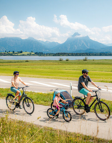 Das Bild fängt eine ruhige Szene von einer Familie mit einem Mann, einer Frau und einem kleinen Kind ein, die Fahrräder auf einem Pfad neben einer Straße fahren. Der Mann hat zudem noch einen Kinderanhänger am Fahrrad, in dem sich noch ein anderes Kleinkind befindet. Der Pfad ist von üppigem Grün umgeben. Im Hintergrund erkennt man den Hopfensee und die Berge. Der Himmel darüber ist ein klarer Blau mit fluffigen weißen Wolken. Die Radfahrer sind in legerer Kleidung und sommerlich angezogen. Sie tragen alle einen Helm. 