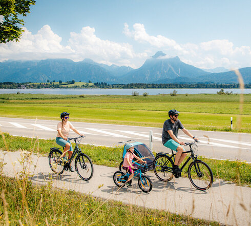 Das Bild fängt eine ruhige Szene von einer Familie mit einem Mann, einer Frau und einem kleinen Kind ein, die Fahrräder auf einem Pfad neben einer Straße fahren. Der Mann hat zudem noch einen Kinderanhänger am Fahrrad, in dem sich noch ein anderes Kleinkind befindet. Der Pfad ist von üppigem Grün umgeben. Im Hintergrund erkennt man den Hopfensee und die Berge. Der Himmel darüber ist ein klarer Blau mit fluffigen weißen Wolken. Die Radfahrer sind in legerer Kleidung und sommerlich angezogen. Sie tragen alle einen Helm. 
