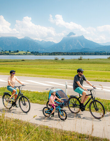 Das Bild fängt eine ruhige Szene von einer Familie mit einem Mann, einer Frau und einem kleinen Kind ein, die Fahrräder auf einem Pfad neben einer Straße fahren. Der Mann hat zudem noch einen Kinderanhänger am Fahrrad, in dem sich noch ein anderes Kleinkind befindet. Der Pfad ist von üppigem Grün umgeben. Im Hintergrund erkennt man den Hopfensee und die Berge. Der Himmel darüber ist ein klarer Blau mit fluffigen weißen Wolken. Die Radfahrer sind in legerer Kleidung und sommerlich angezogen. Sie tragen alle einen Helm. 