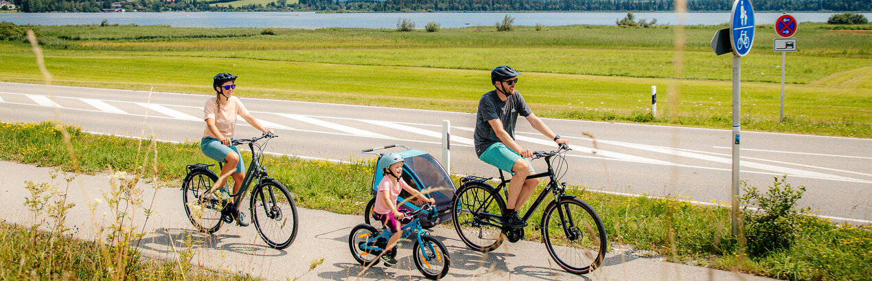 Das Bild fängt eine ruhige Szene von einer Familie mit einem Mann, einer Frau und einem kleinen Kind ein, die Fahrräder auf einem Pfad neben einer Straße fahren. Der Mann hat zudem noch einen Kinderanhänger am Fahrrad, in dem sich noch ein anderes Kleinkind befindet. Der Pfad ist von üppigem Grün umgeben. Im Hintergrund erkennt man den Hopfensee und die Berge. Der Himmel darüber ist ein klarer Blau mit fluffigen weißen Wolken. Die Radfahrer sind in legerer Kleidung und sommerlich angezogen. Sie tragen alle einen Helm. 