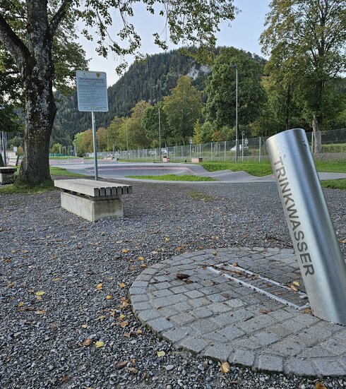Das Bild zeigt einen Trinkbrunnen bestehend aus einem langen Metallrohr auf dem „Trinkwasser“ steht. Dahinter liegen die Parcours des Skate- und Bikeparks. Dazwischen erheben sich hohen Bäume. Im Hintergrund sind bewaldete Hügel zu sehen.   