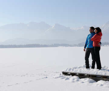 Das Bild zeigt einen Mann und eine Frau. Der Mann hat den Arm um die Frau gelegt. Sie stehen auf einem verschneiten Holzsteg. Der davorliegende See ist tief verschneit und nicht als See zu erkennen. Im Hintergrund erheben sich die schneebedeckten Berge. Die Landschaft ringsum ist tief verschneit. Der Himmel ist blau. Die Sonne scheint. 
