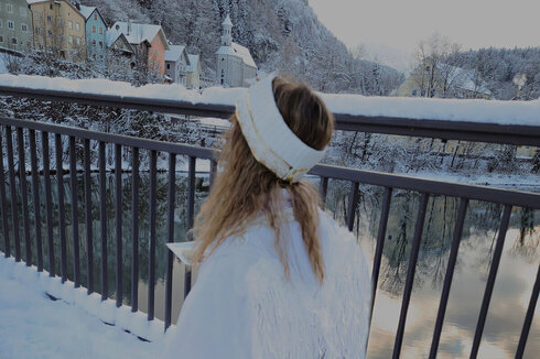 Das Bild fängt eine idyllische Winterlandschaft ein. Eine junge Frau, angekleidet in ein weißes Kleid und einen passenden weißen Hut, ist die Hauptperson des Fotos. Sie ist dabei, auf einem schneebedeckten Bürgersteig zu gehen, ihre roten Schuhe kontrastieren mit dem weißen Schnee. Die Frau sieht nach unten, vielleicht auf den Schnee unter ihren Füßen.  Im Hintergrund gibt es eine schöne Aussicht auf einen Fluss, über den eine Brücke spannt. Die Brücke ist mit Schnee verziert, was zur wintersportlichen Atmosphäre hinzufügt. Auf der anderen Seite des Flusses gibt es bunte Häuser, deren leuchtende Farben sich gegen den weißen Schnee abheben.  Der Himmel über ist bedeckt, was ein sanftes Licht über die gesamte Szene wirft. Das Bild ist eine wunderschöne Darstellung eines Wintertages, mit der Reise der Frau entlang des schneebedeckten Bürgersteiges als Höhepunkt