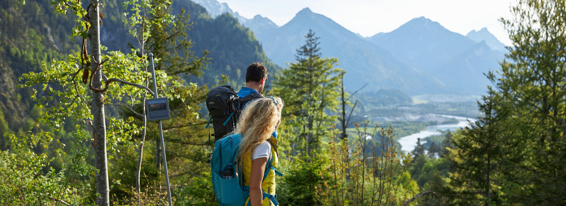 Das Bild zeigt einen Mann und eine Frau in Wanderkleidung und mit Rucksäcken, die auf einer Anhöhe stehen und ins Tal blicken, durch das ein Fluss fliest. Im Hintergrund erheben sich die Berge. Ringsum wachsen Bäume und Büsche. Der Himmel ist blau. Die Sonne scheint. 