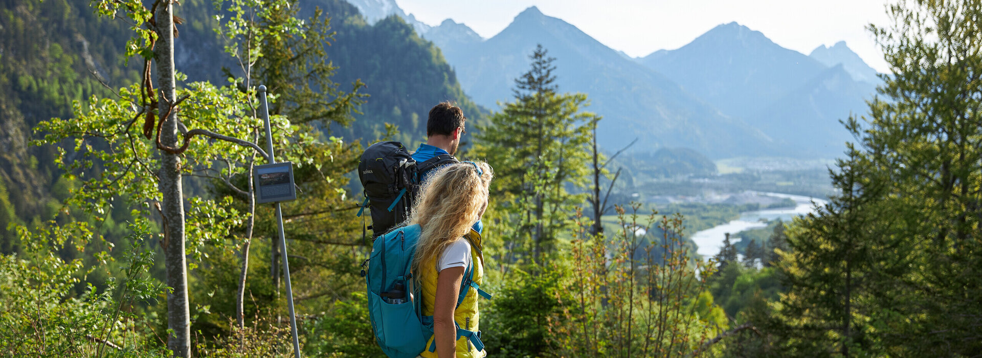 Das Bild zeigt einen Mann und eine Frau in Wanderkleidung und mit Rucksäcken, die auf einer Anhöhe stehen und ins Tal blicken, durch das ein Fluss fliest. Im Hintergrund erheben sich die Berge. Ringsum wachsen Bäume und Büsche. Der Himmel ist blau. Die Sonne scheint. 