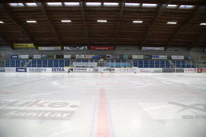 Man blickt von der Eisfläche eines Eisstadions auf die leere Zuschauertribüne. Die Decke des Stadions ist mit Lampen hell erleuchtet. An der Bande, den Wänden und unter der Eisfläche sind Werbeschilder zu sehen. Das Stadion ist menschenleer.   