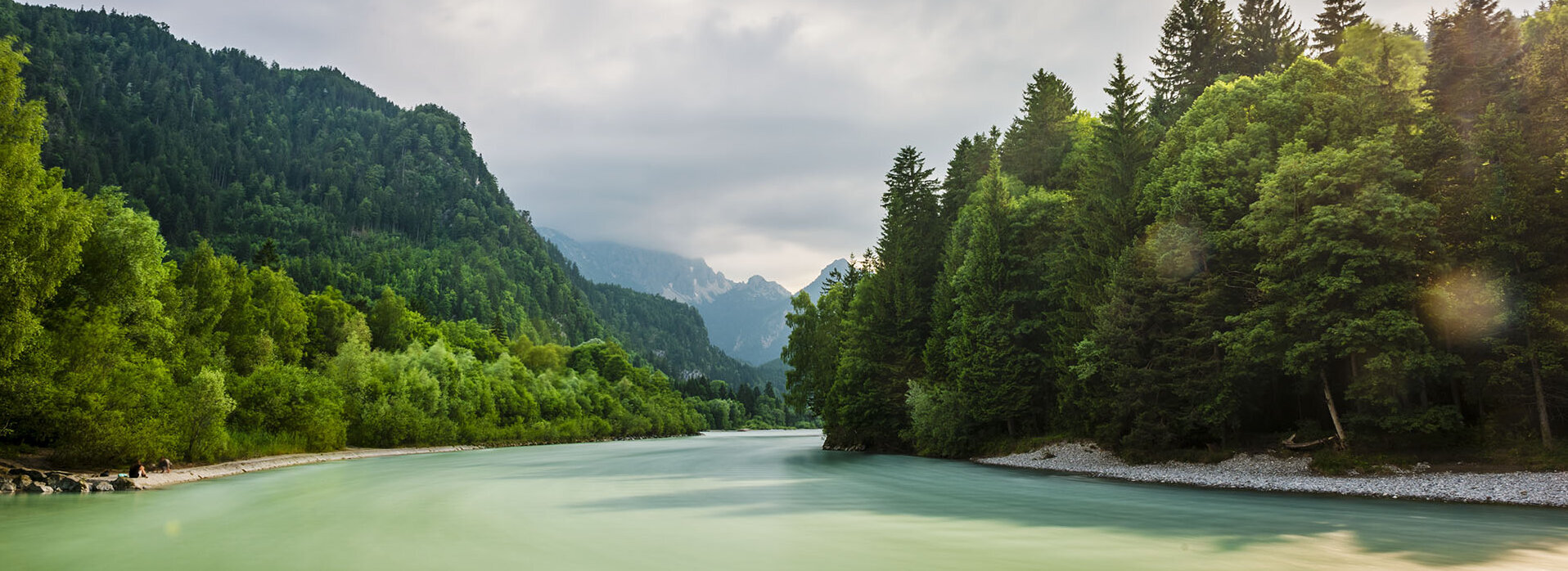 Das Bild zeigt eine friedliche Landschaft um den Fluss Lech, der durch einen grünen Wald fließt. Der Fluss, mit seinem leicht türkisenen Wasser, ist der zentrale Fokus des Bildes. Er windet sich durch den dichten Wald, erzeugt einen Sinn von Ruhe und Harmonie mit der Natur. Der Wald ist ein kräftiges Grün, gefüllt mit hohen Bäumen, die über den Fluss hineinwachsen. Der blaue Himmel darüber ist mit flauschigen weißen Wolken durchzogen, die zur Schönheit der Szene beitragen. Das Bild wird aus der Ferne aufgenommen, was einen umfassenden Blick auf den Fluss sowie den umliegenden Wald erlaubt. Die Perspektive des Bildes betont Tiefe, was die Weite des Waldes und des Flusses unterstreicht. Das Bild ist eine schöne Darstellung von Naturfrieden und der Harmonie zwischen dem Fluss und dem Wald.