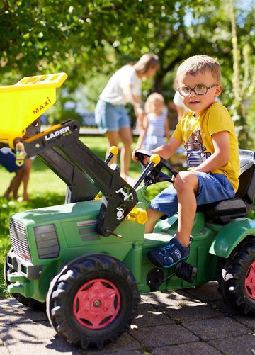 Das Bild zeigt einen jungen Jungen, der in einem gelben Oberteil und blauen Shorts sitzt, auf einem grünen und gelben Rasenmäher. Der Mäher ist mit einem gelben Eimer am Hinterteil ausgestattet. Der Junge ist aktiv in den Mähvorgang involviert, indem er sich an den Lenkerhängen fasst und den Mäher manövriert. Im Hintergrund sind Bäume und ein Haus zu sehen, was auf eine ländliche Umgebung hindeutet. Die Entschlossenheit und Konzentration des Jungen sind offensichtlich, wie er den Mäher bedient. Das Bild strahlt ein Gefühl von kindlicher Neugier und Abenteuerlust aus.